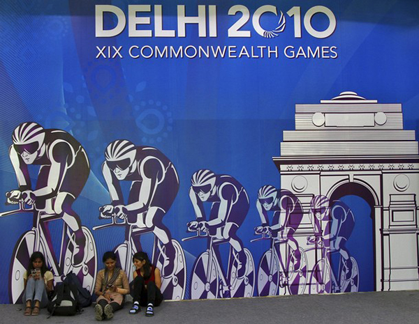 An official billboard for Delhi 2010 with dark blue background shows a team of cyclists riding through an arc