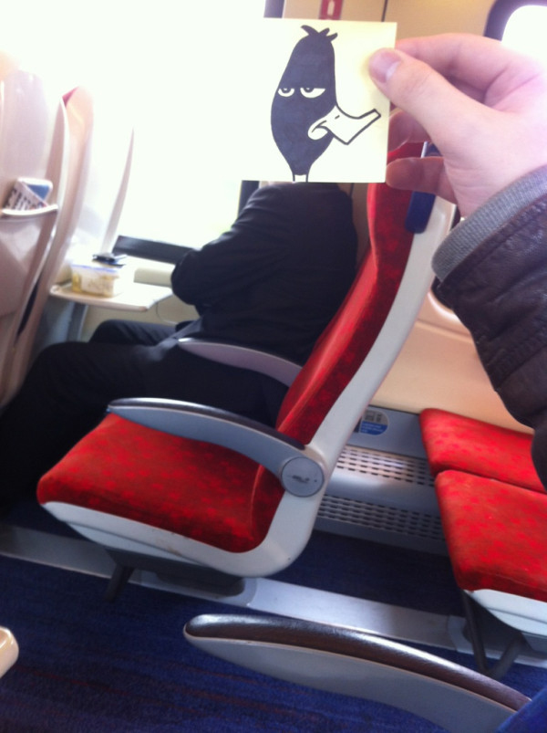 Funny sketch of a duck's face set to the side as if it had been shot. October Jones has put the sticky note drawing on a commuter's body on the train