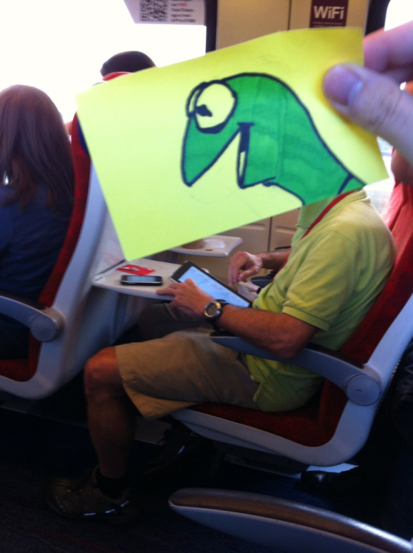 Funny sketch of Kermit The Frog smiling and looking really happy superimposed onto a real life person's head on the train to work looking at his iPad wearing a green polo.