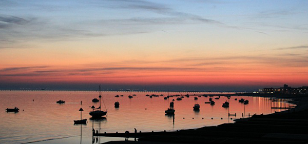 Gorgeous sunset with pinks, peaches, blue and oranges. The boats and land are in a black silhouette against the water which reflects the perfect setting of the sun