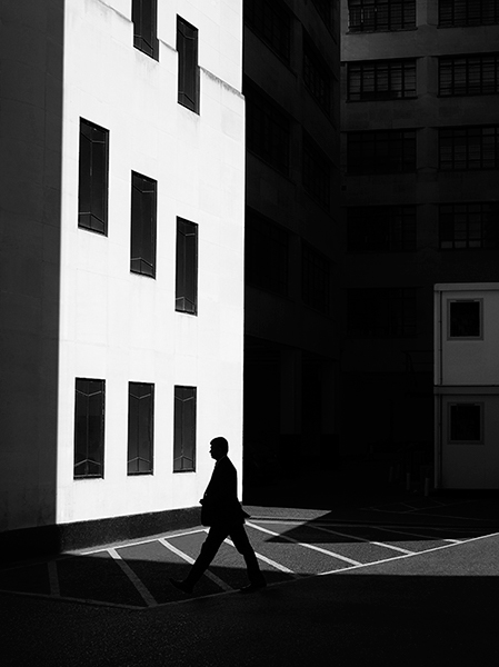 Black and white, light and shadow effect photograph with a bright-white building with blackened out windows and a black, silhouette of a man walking past