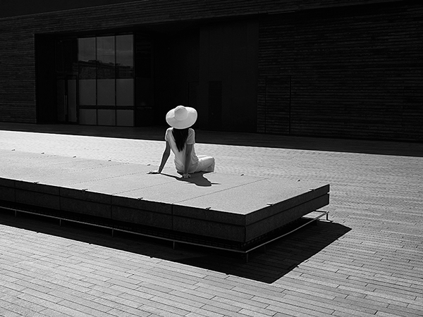 Light and shadow, black and white photograph showing a woman perched on a rectangular shaped , long bench with her back facing us. She is wearing a white hate and white t-shirt and trousers, looking at a dark, block building . The floor is made of wood.