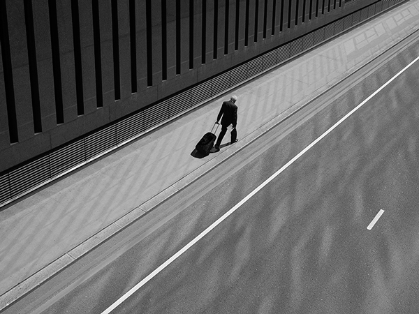 Black and white photograph taken overhead and looking down on the subject. Subject is wearing a black suit, trailing behind him a black suitcase / work bag. He is walking along a silvery path next to a road where the building's windows from next to him is reflecting on the road.